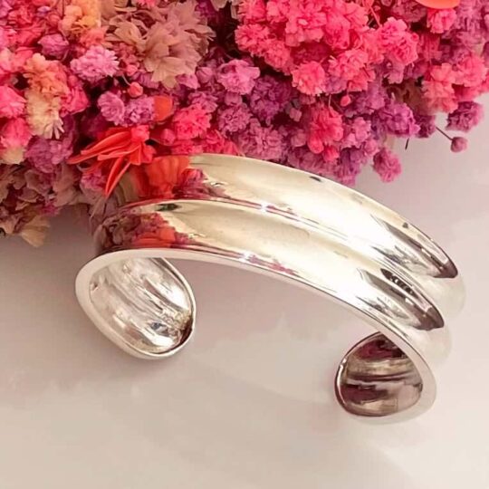 Shiny silver cuff bracelet with a modern, open design, placed beside vibrant pink flowers. Perfect for stylish accessories and gift ideas, enhancing any outfit with elegance.