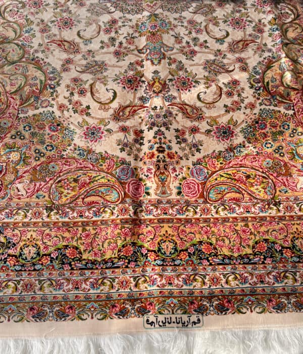 An upclose look at a silk persian rug with detailed floral and wildlife patterns and the makers mark in Arabic. The shade is cream and pink.